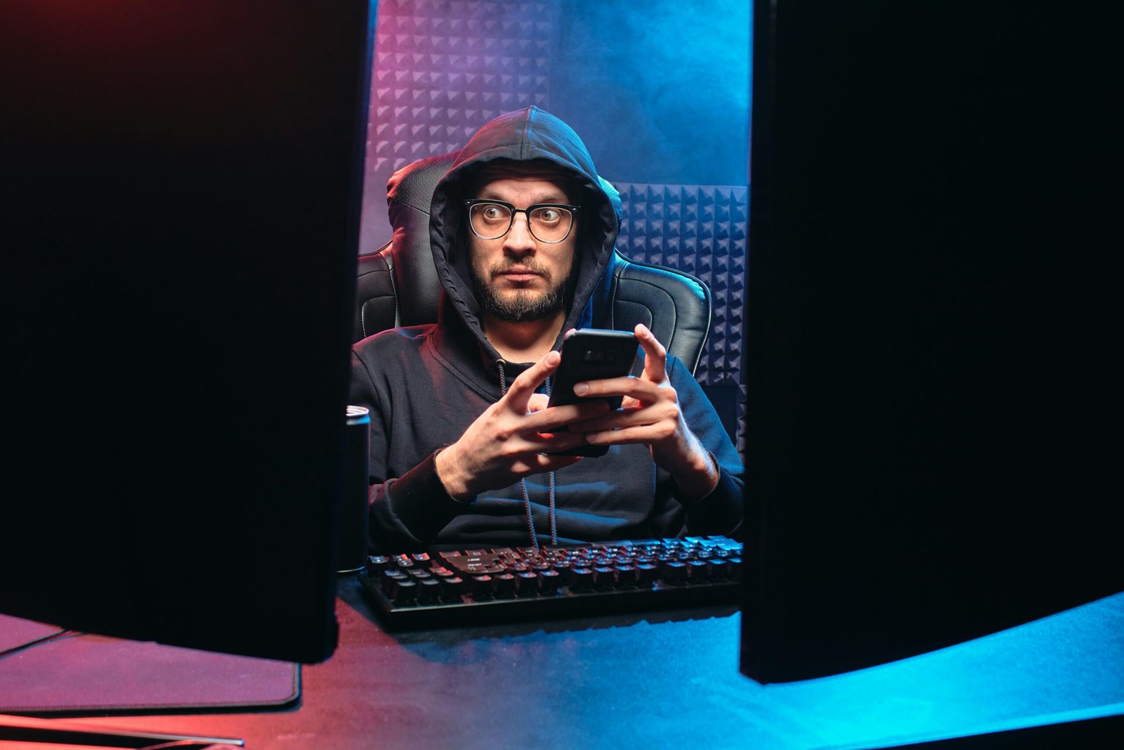 Man in a hoodie holding a phone while sitting between two computer screens, symbolizing cybersecurity.